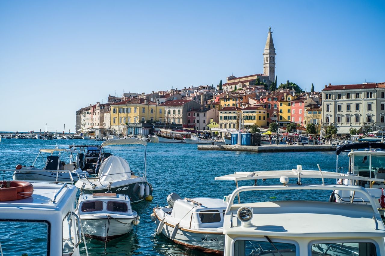 Der Hafen von Rijeka, mit vielen Booten im Vordergrund, die im klaren blauen Wasser schwimmen. Im Hintergrund ist die historische Altstadt von Rijeka zu sehen, mit bunten Häusern und der markanten Kirche, die auf einem Hügel thront.