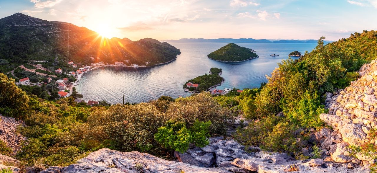 Ein atemberaubender Blick auf die Insel Mljet bei Sonnenuntergang. Die Sonne geht hinter den bewaldeten Hügeln unter und taucht die Bucht und das Dorf in goldenes Licht. Das ruhige Wasser der Bucht spiegelt den Himmel wider, und kleine Inseln sind im Hintergrund zu sehen, umgeben von üppigem Grün und felsigen Hängen.